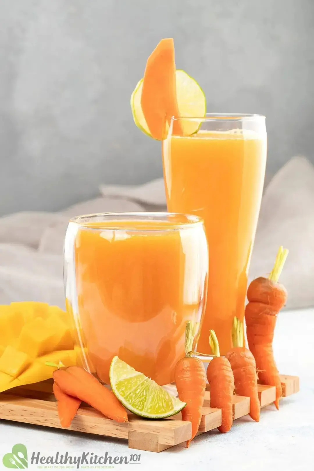 Two glasses of carrot smoothie, one tall and one short, put next to a lime wedge and many baby carrots