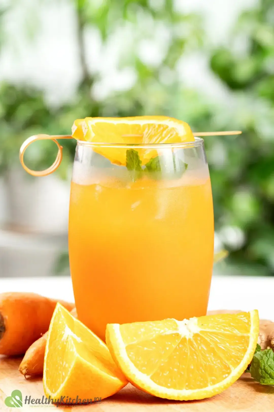 A glass of orange carrot juice garnished with orange wedges and carrots, with a blurred greeneries background