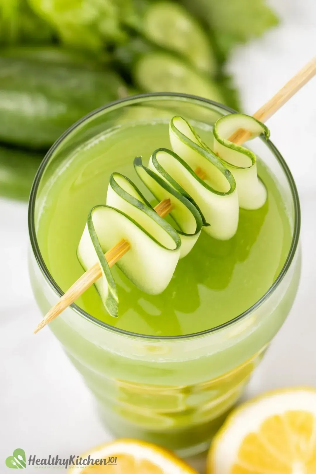 A glass of cucumber celery drink topped with a skewered cucumber slice and next to some lemons
