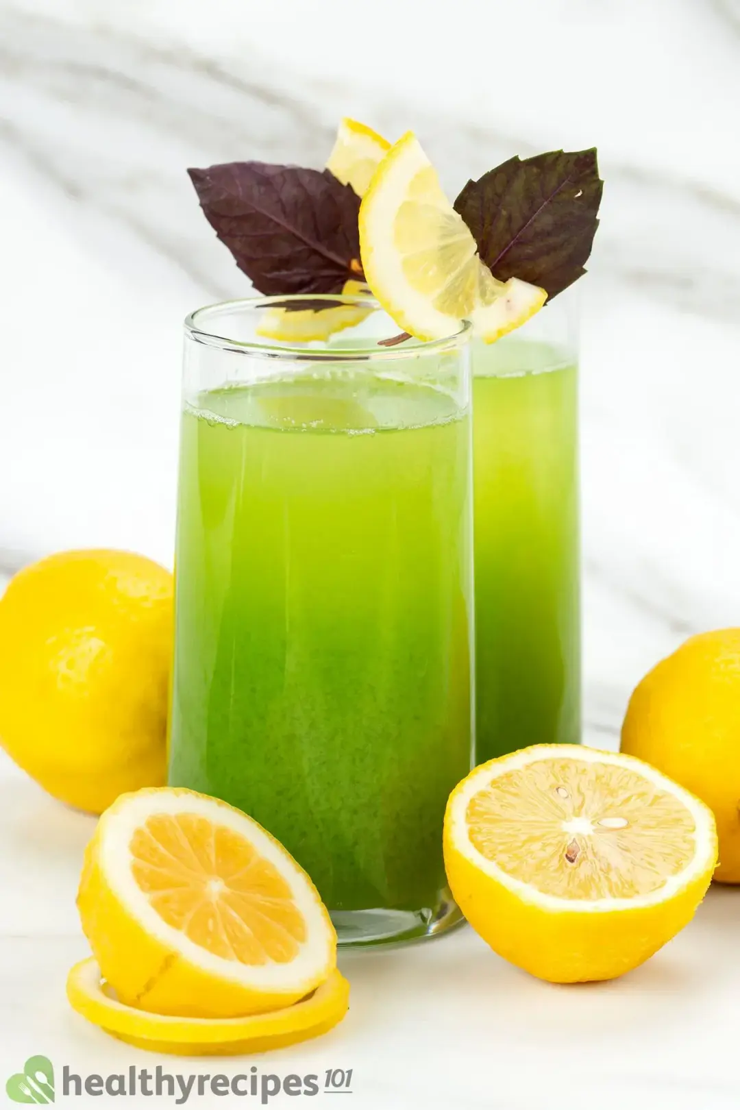 Two tall glasses of cucumber and lemon juice put next to lemon wheels and topped with purple leaves