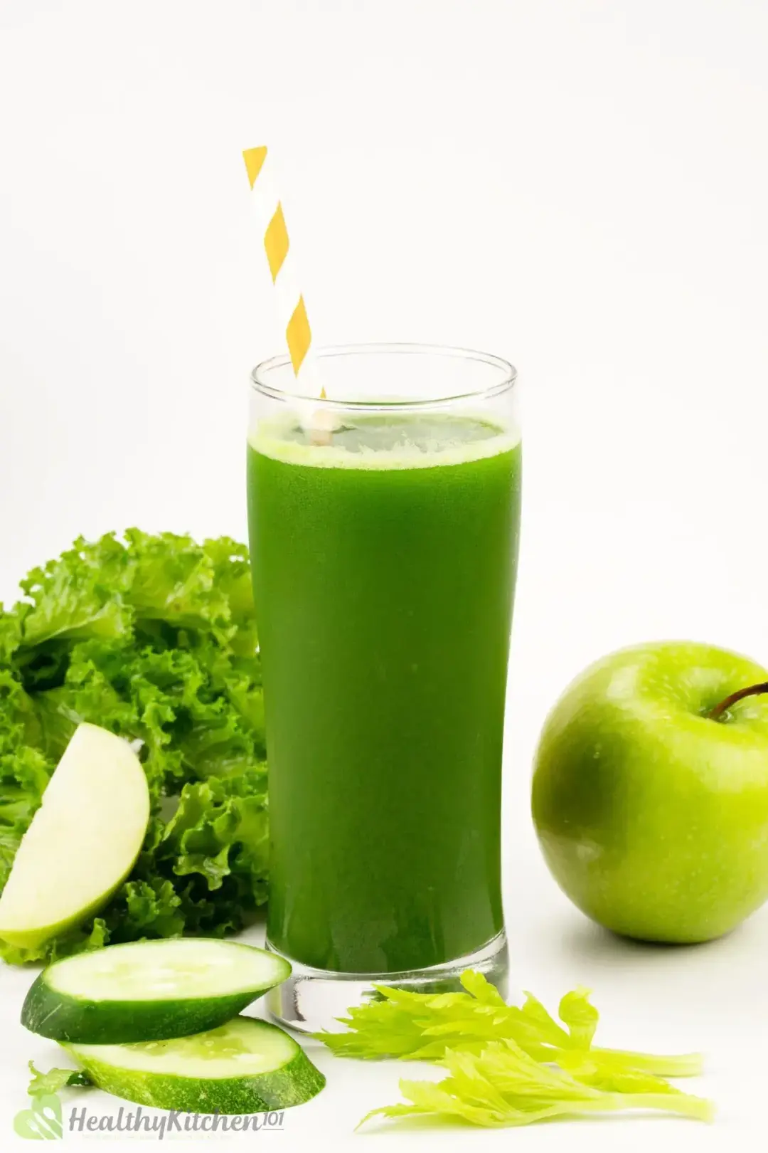 A cool glass of green vegetable juice, with an orange-striped straw centering decorants: a whole green apple, thick slices of cucumber, and some green leaves