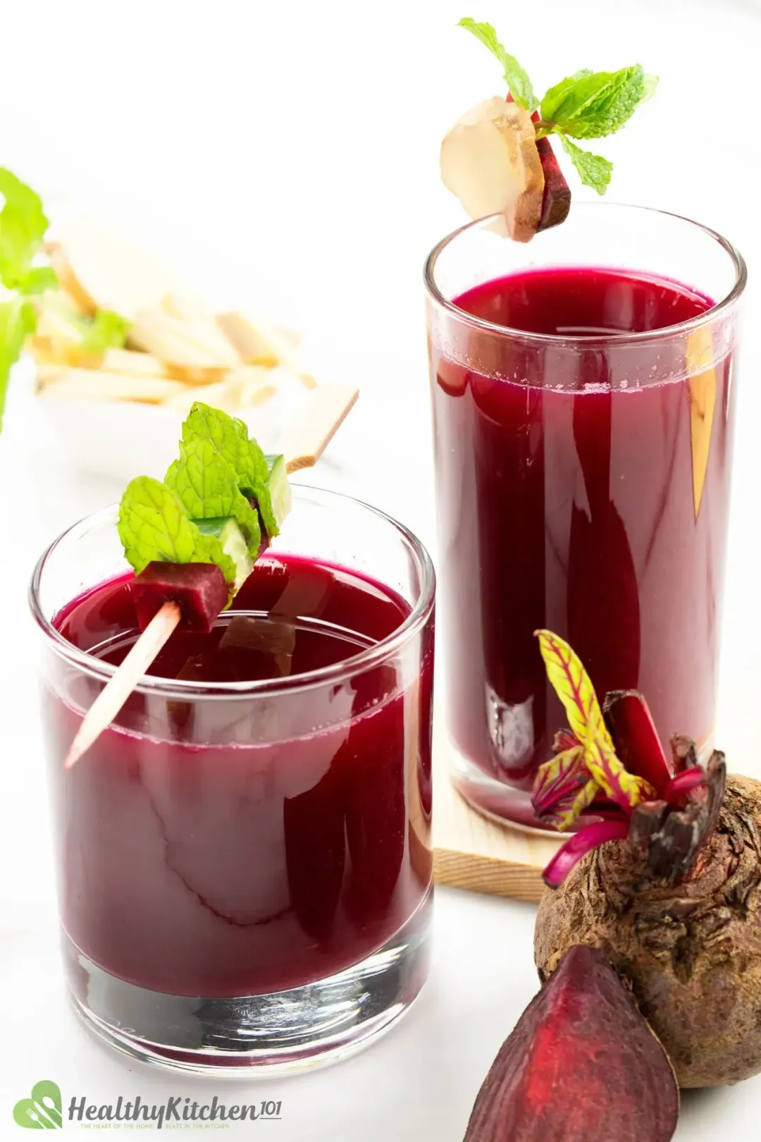 One short glass and one tall glass of beetroot juice, each topped with mints, ginger pieces, and beetroot pieces next to some whole beets