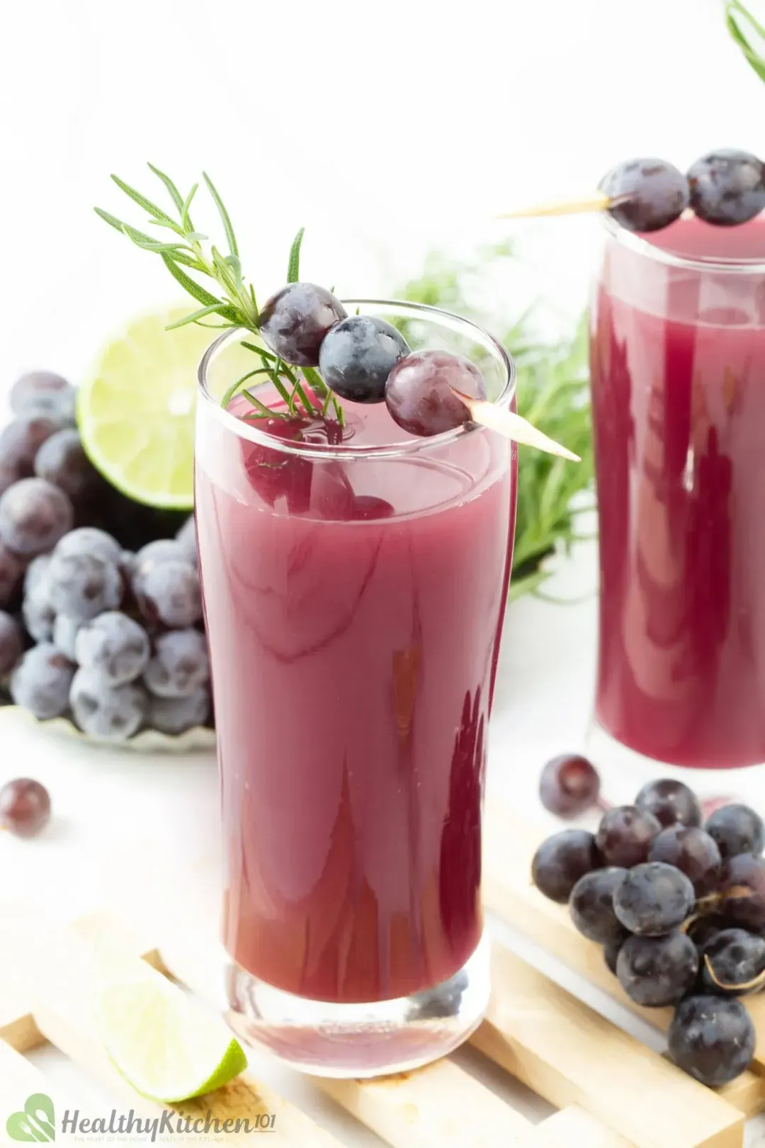 A tall glass of purple grape juice with a rosemary sprig, lime, rosemary, and grapes all around