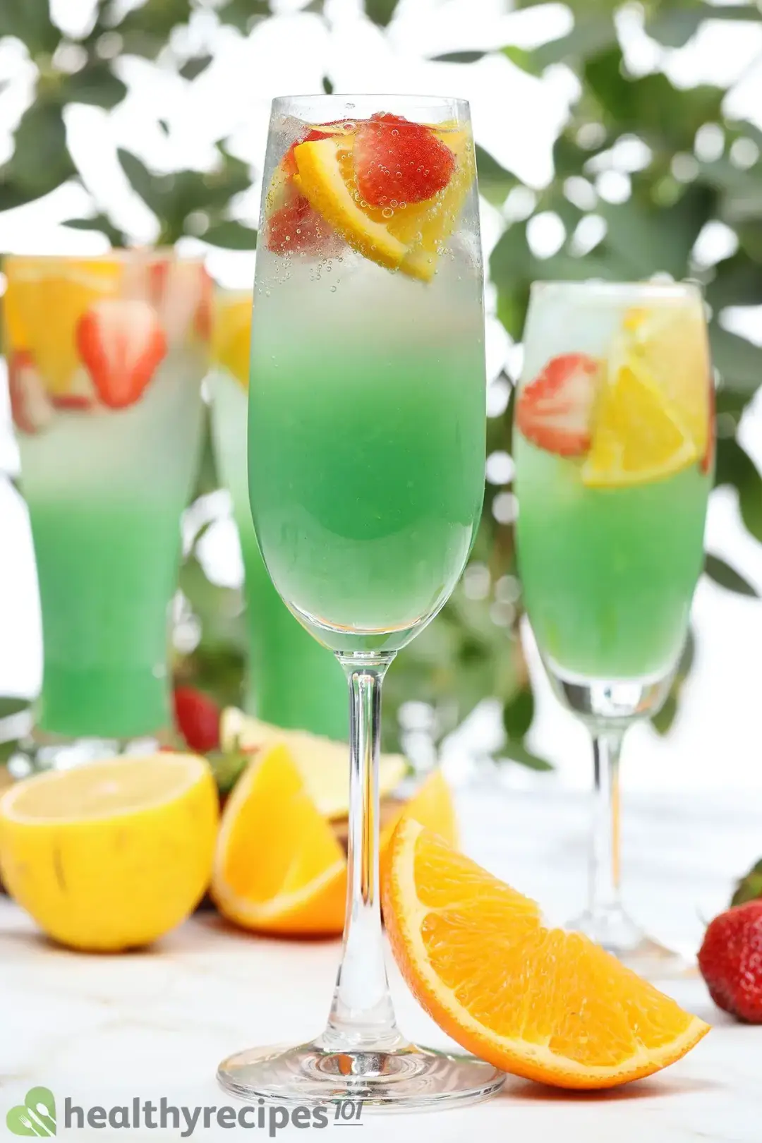 Multiple glasses of green jungle juice surrounded by lemon slices, orange slices, and strawberry slices