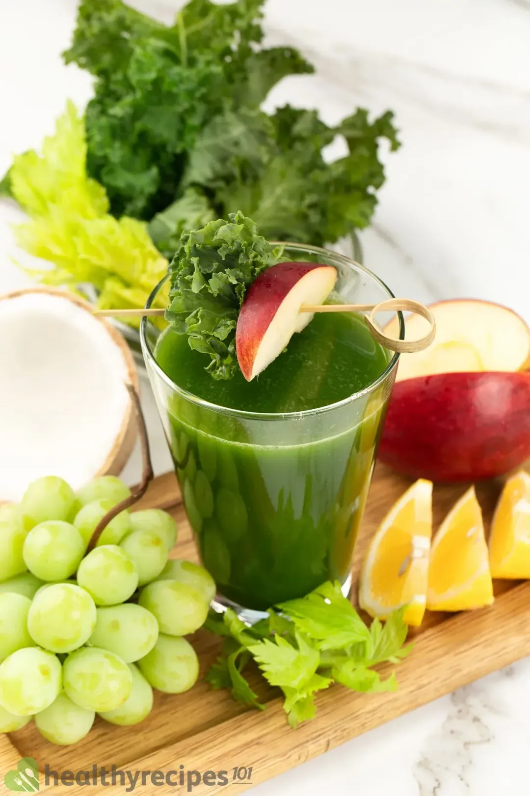 A glass of green machine juice, some grapes, celery, three wedges of lemon, and an apple placed on a long wooden board with some greens on the background