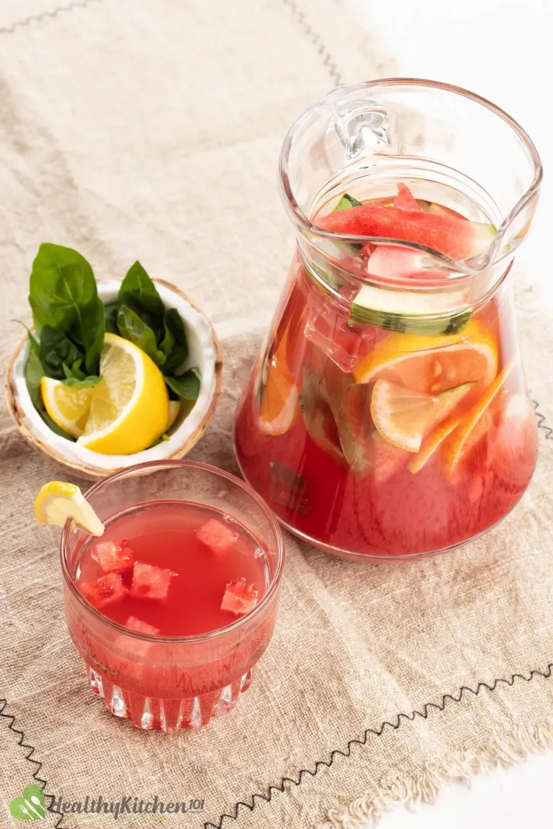 A close up picture of jug and a glass of watermelon jungle juice placed on a rug next to a coconut shell with lemon slices and basil leaves in it.