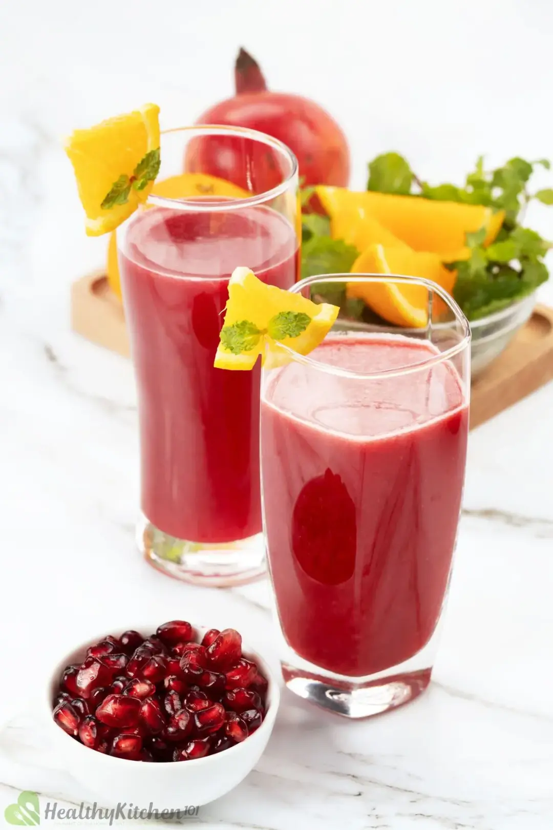 Two glasses of orange pomegranate juice garnished with orange wedges and put next to a bowl of pomegranate gems
