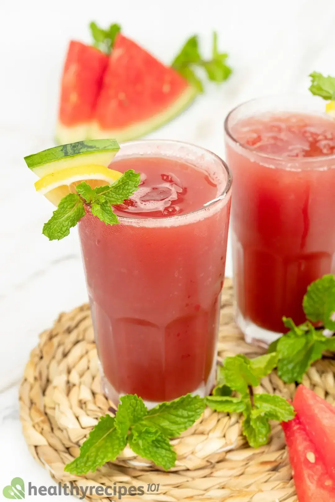 Two dark red glasses of watermelon cucumber juice placed on a woven plate surrounded by spearmint leaves and a few watermelon slices