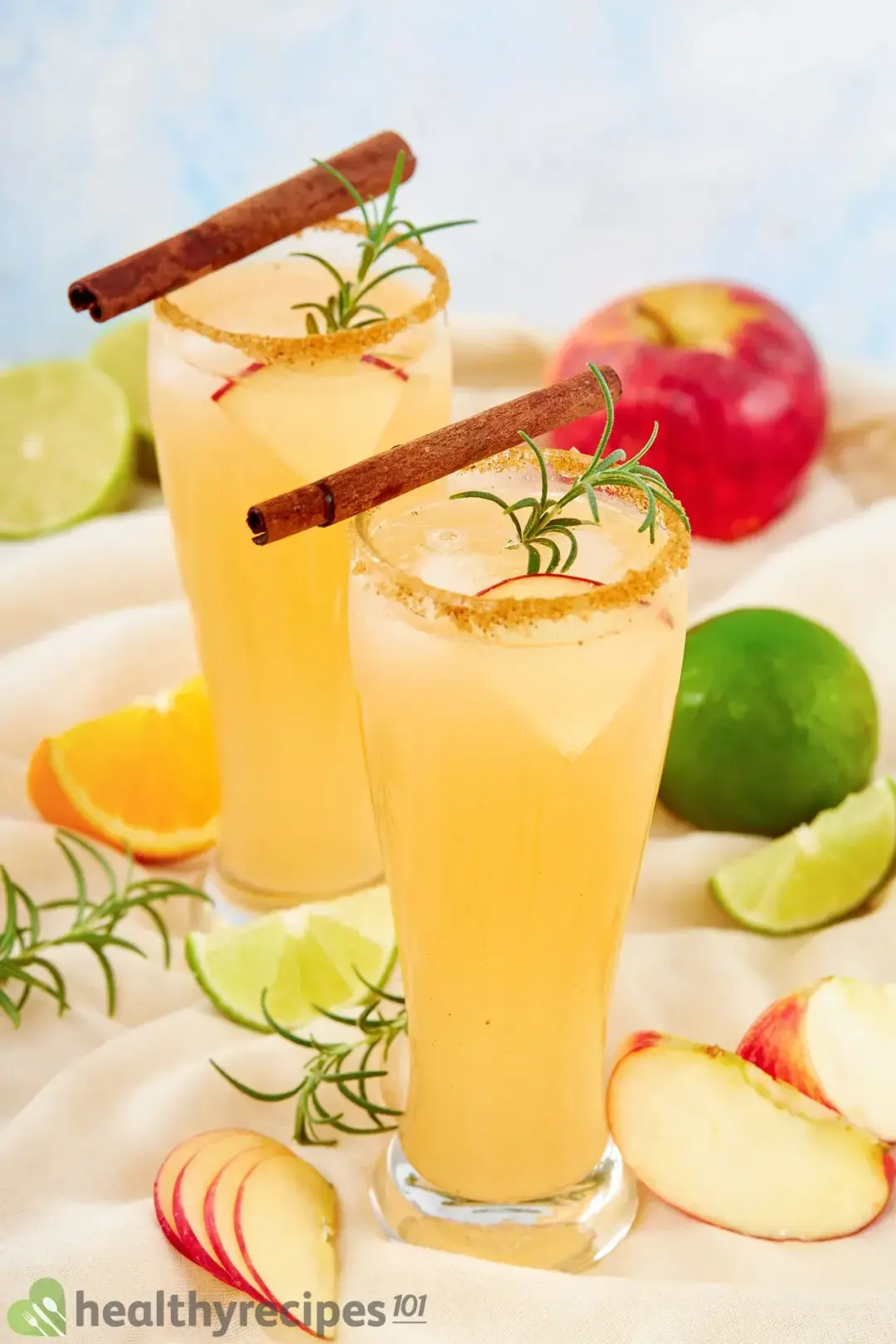 Two glasses of apple cider margarita cocktail topped with cinnamon sticks, rosemary sprigs, and apple and citrus wedges all around