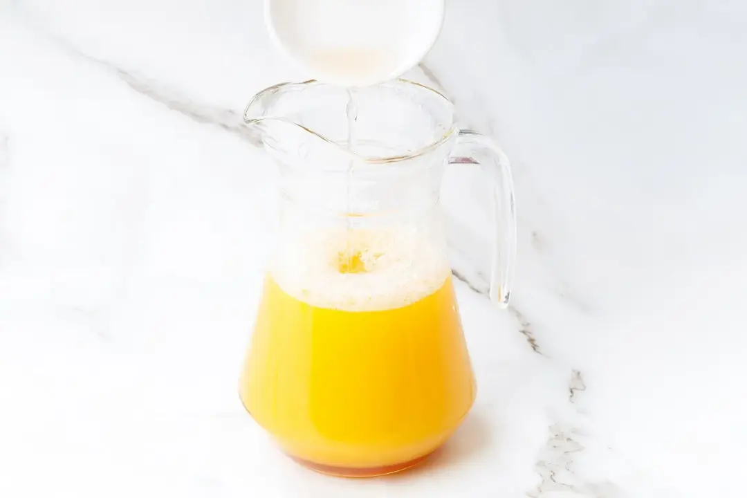 A white bowl pouring some honey into a clear glass pitcher of pineapple juice and cider mixture
