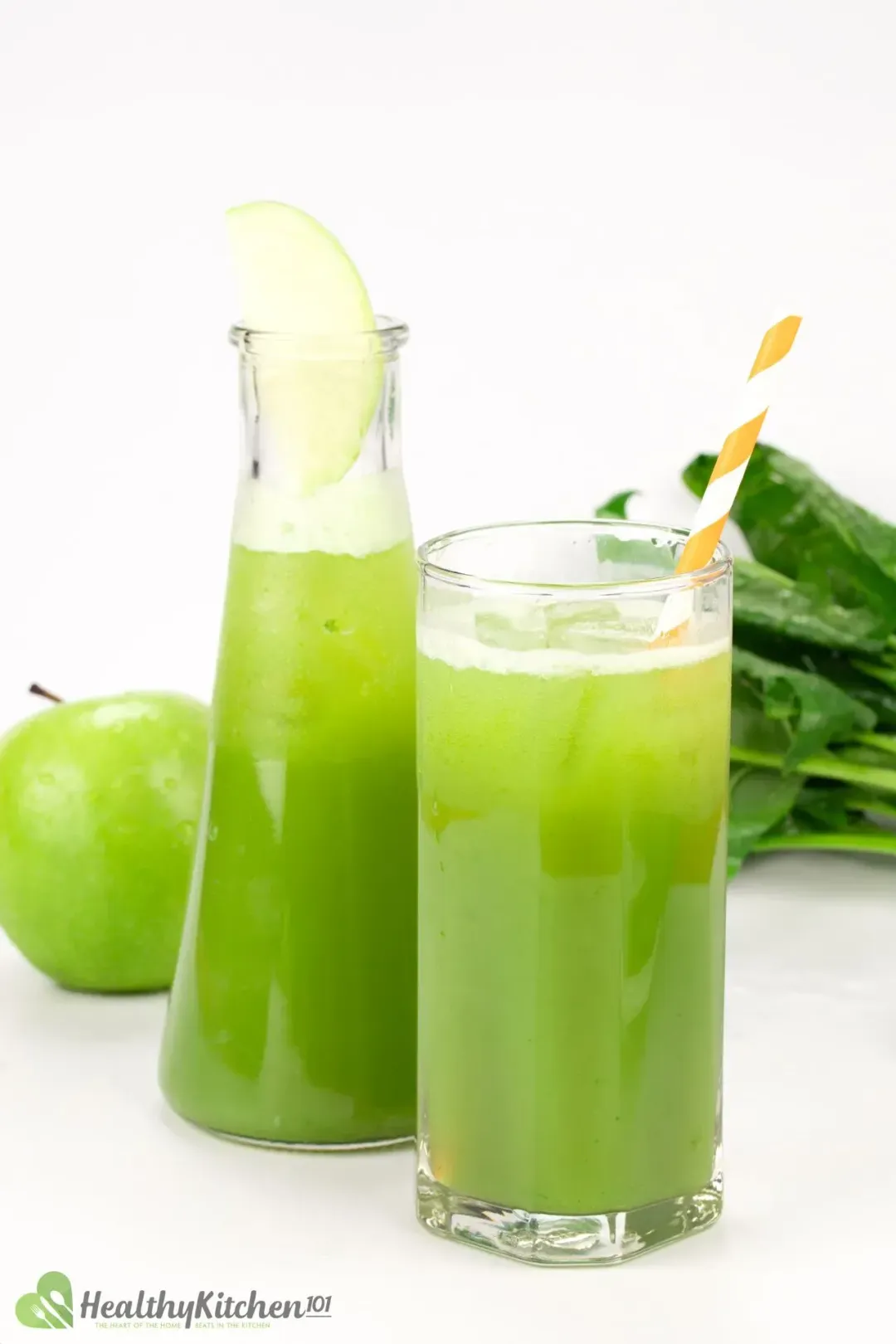 A glass and a pitcher of green apple juice decorated with an orange-striped straw and a slice of green apple with a whole green apple and spinach in the background