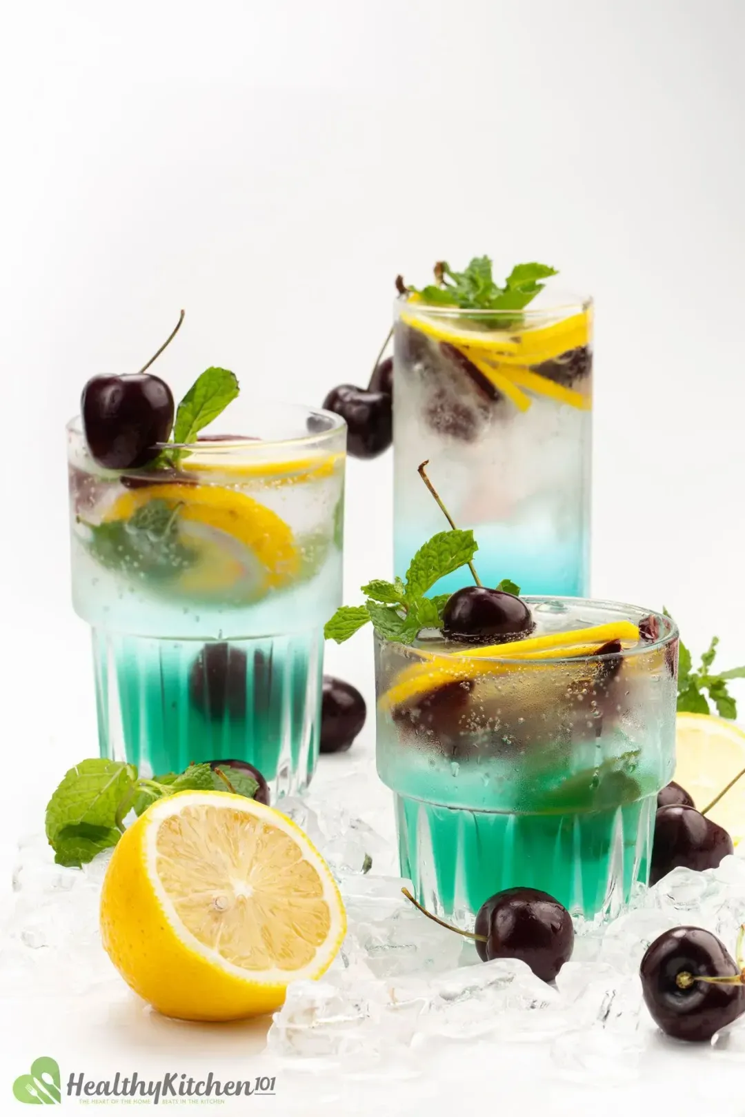 Three cocktail glasses of a cyan-hued drink filled with cherries, lemon wedges, and mints