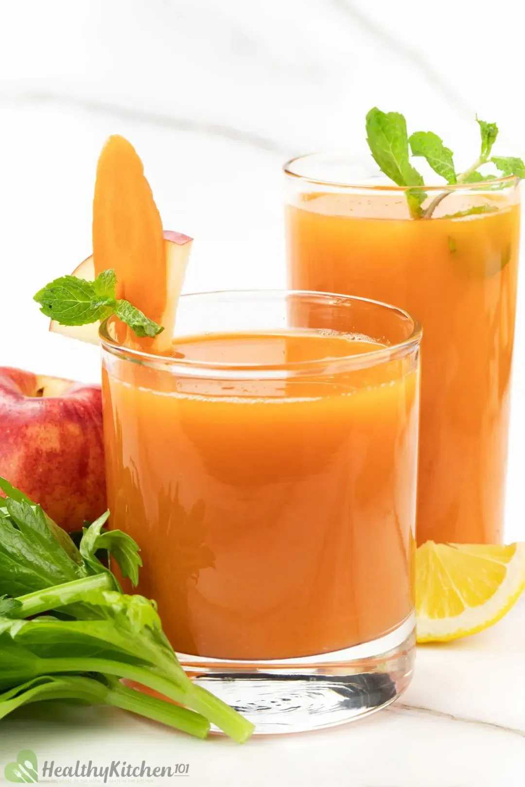 Two short glasses of carrot celery juice garnished with some celery leaves, a carrot slice, next to an apple