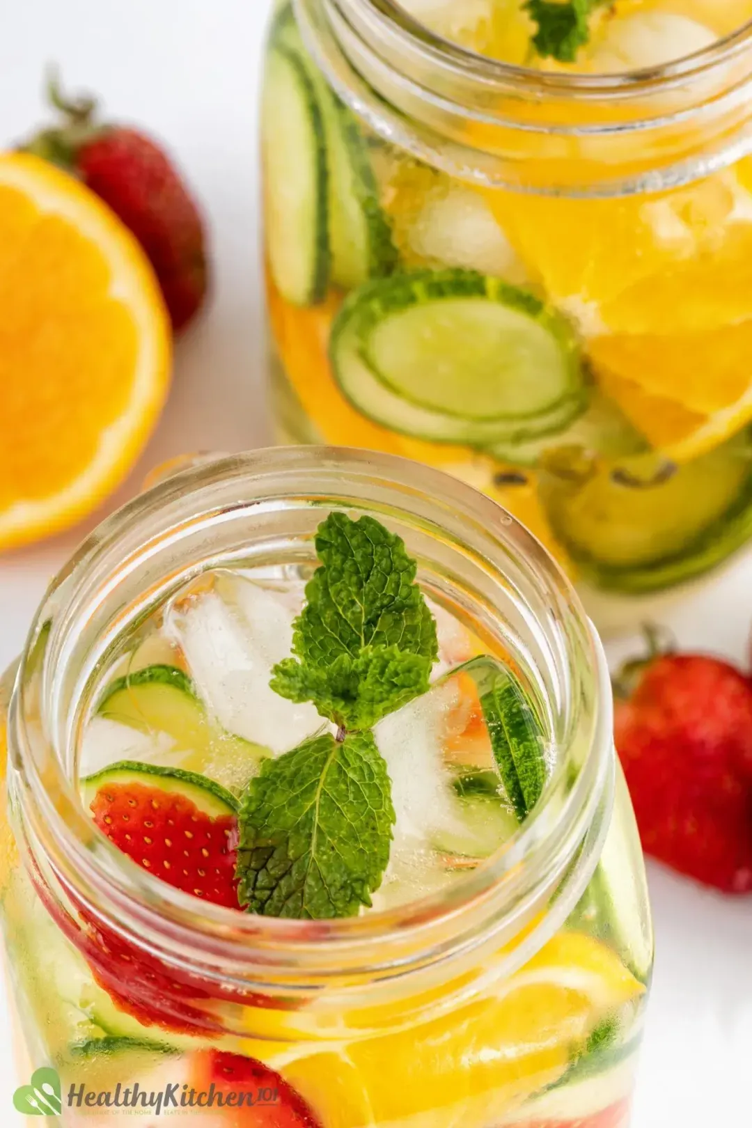 Two jars of clear water submerging strawberries, orange, mint leaves with ice