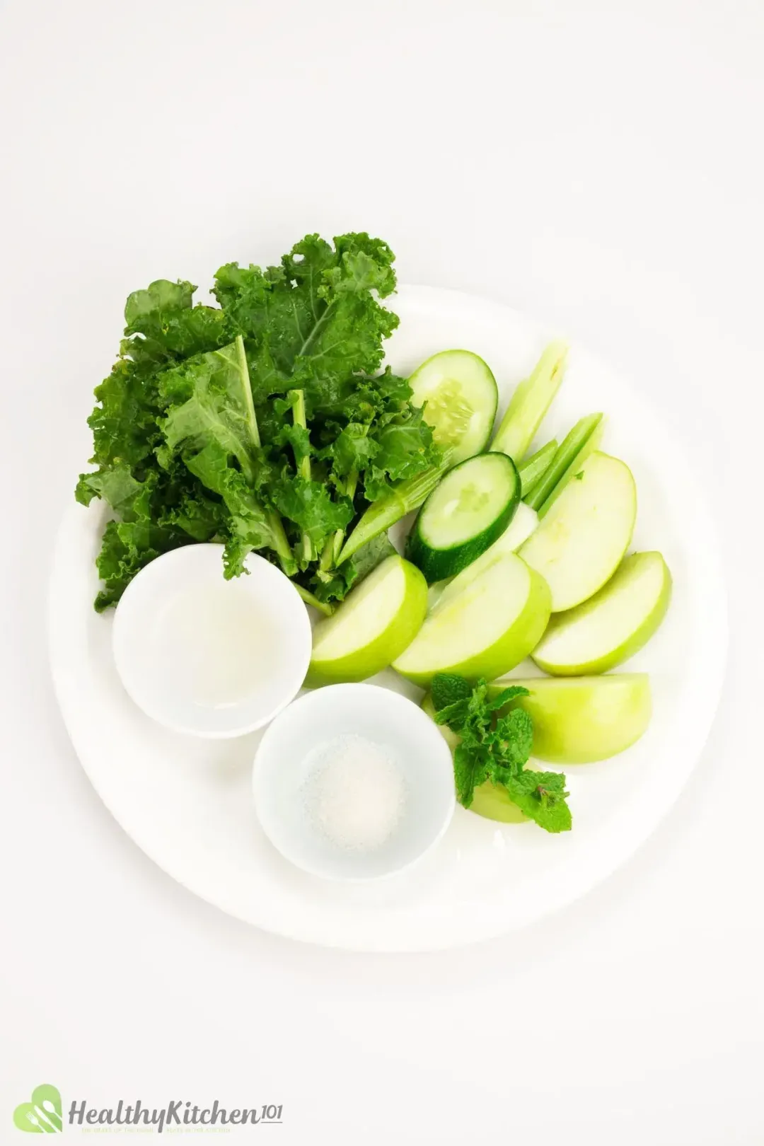 Ingredients of green vegetable juice: kale leaves, celery stalks cut small, cucumber slices, green apple wedges, sugar, lemon juice all placed on a white dish