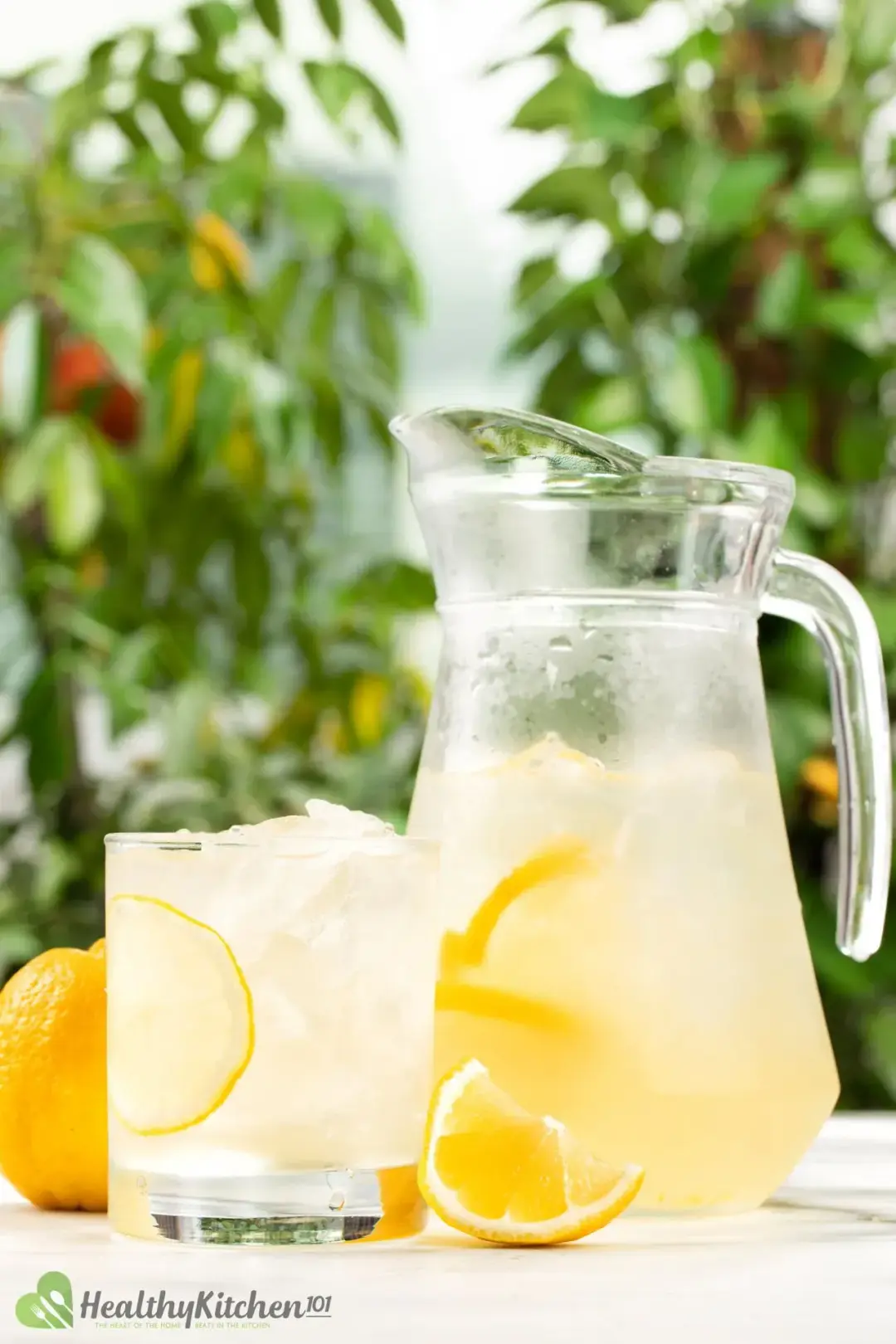  Iced lemonade in a short glass and a pitcher sitting side by side in front of some trees