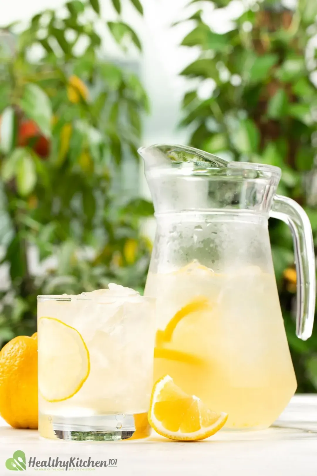 Iced lemonade in a short glass and a pitcher sitting side by side in front of some trees