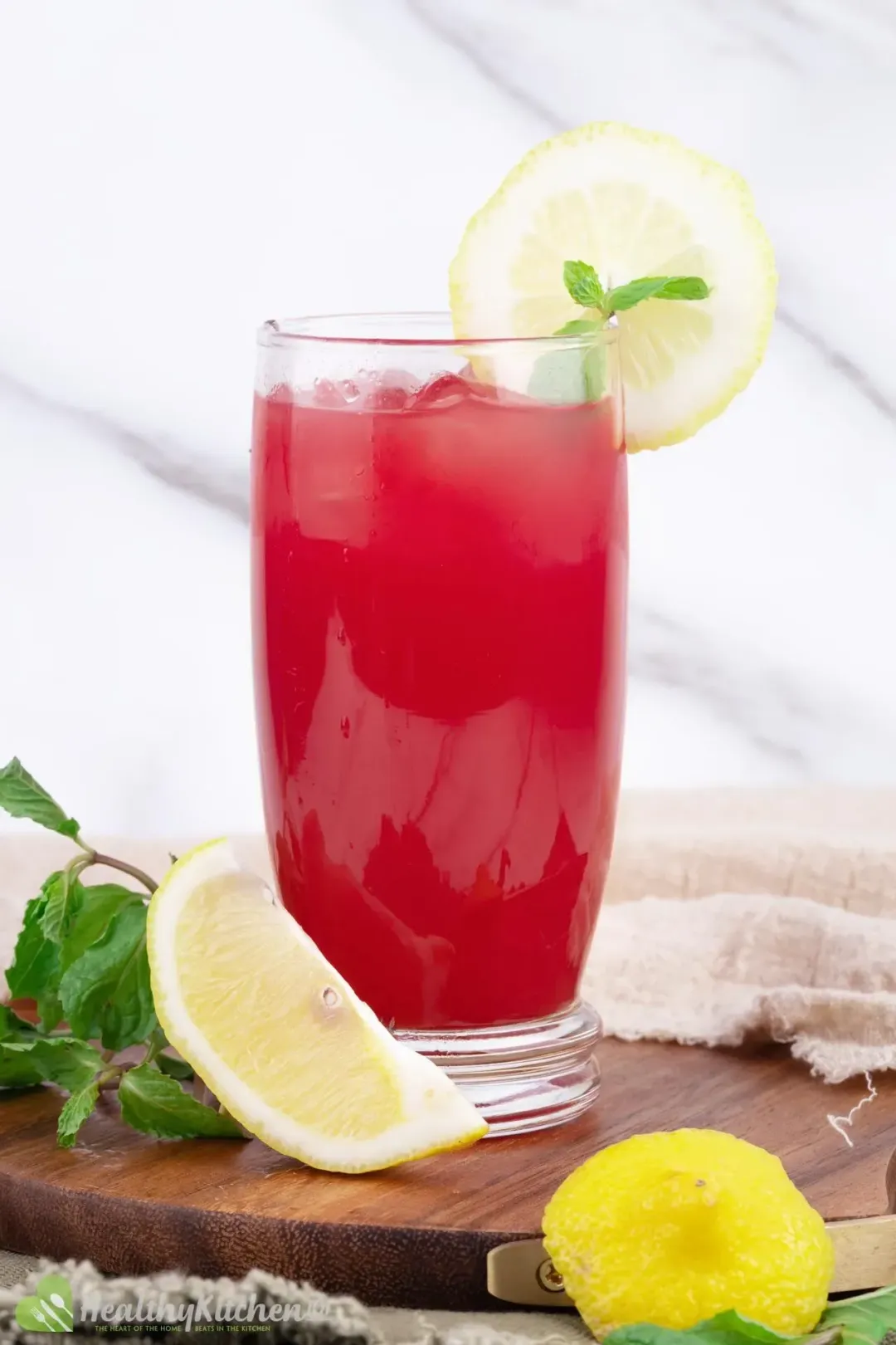 A glass of watermelon juice and lemon with a slice of lemon and a mint leaf placed on the rim surrounded by some fresh mint leaves and wedges of lemon on a wooden tray.