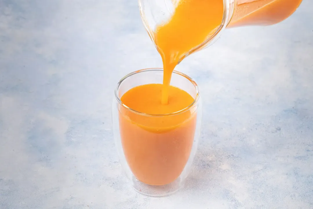 A carrot smoothie poured from a glass pitcher into a smoothie glass