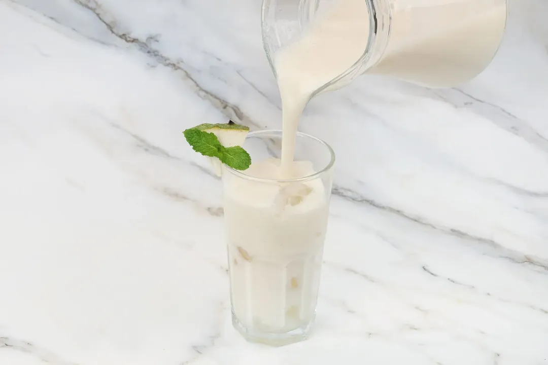 A pitcher of soursop juice pouring into a tall glass, garnished with mints and soursop wedge