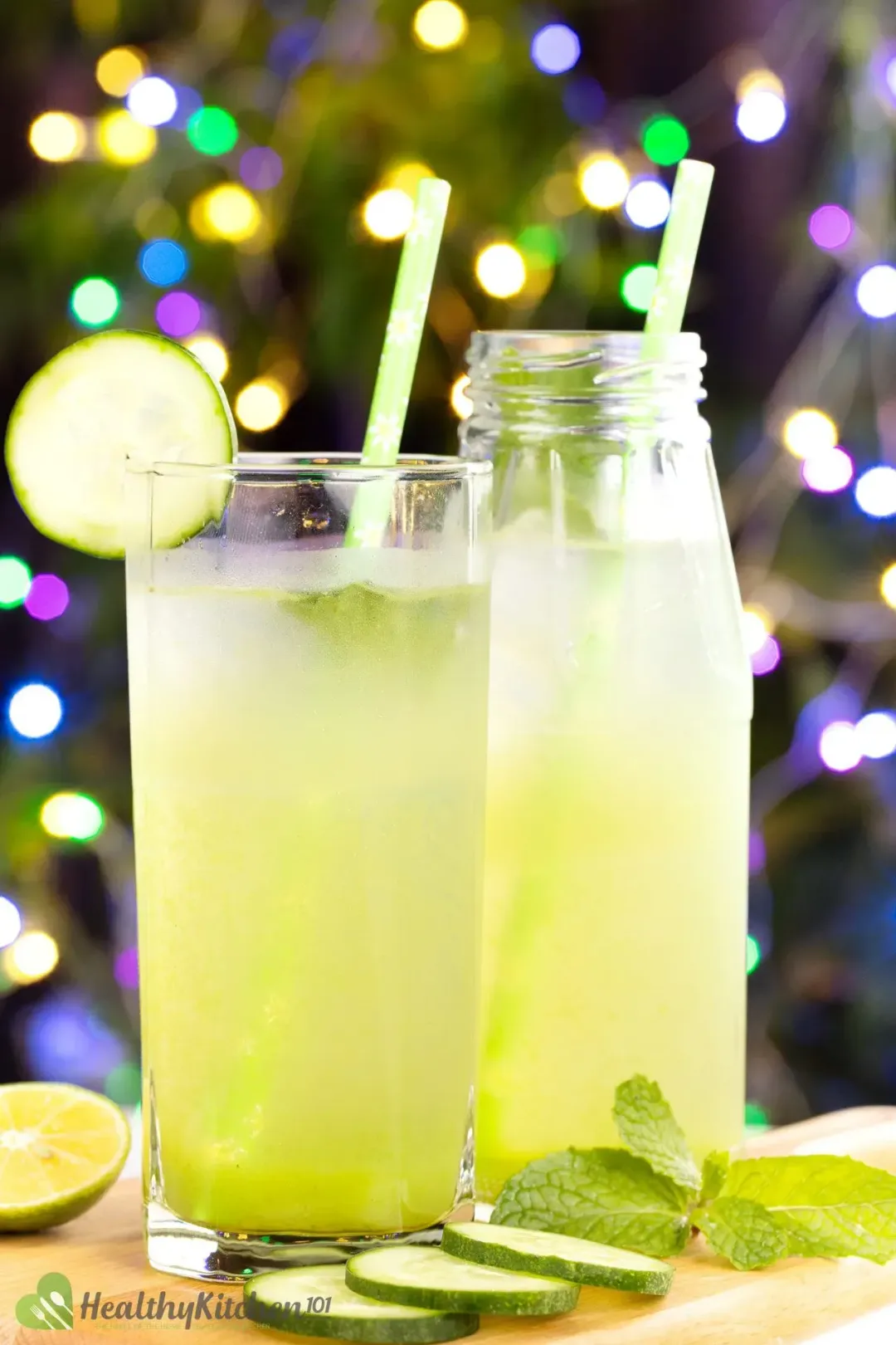 In front of Christmas trees with lights a glass of cucumber and lime juice next to some mints leaves, cucumbers slices, and a jar of drink