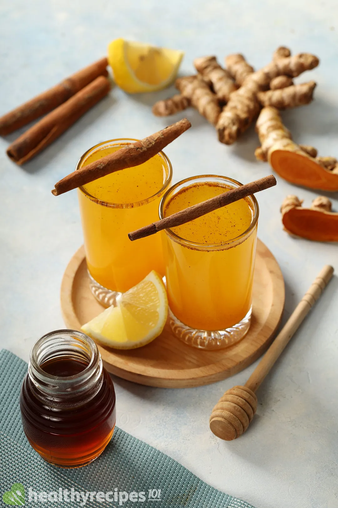 two glasses of yellow liquid next to a honey jar on a table