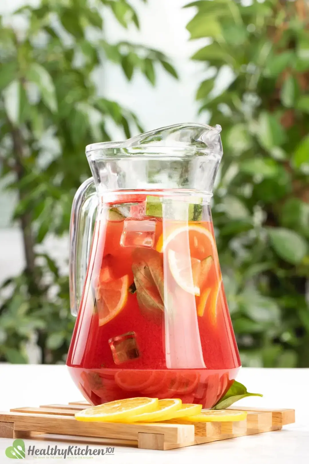 A jug of watermelon jungle juice placed on a wooden board with some lemon slices and basil leaves on the sides