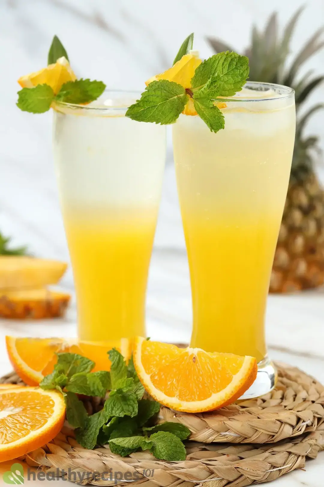 Two glasses of unstirred rum and orange juice soda, put next to garnishes: orange wedges, mint sprigs, and twined coasters