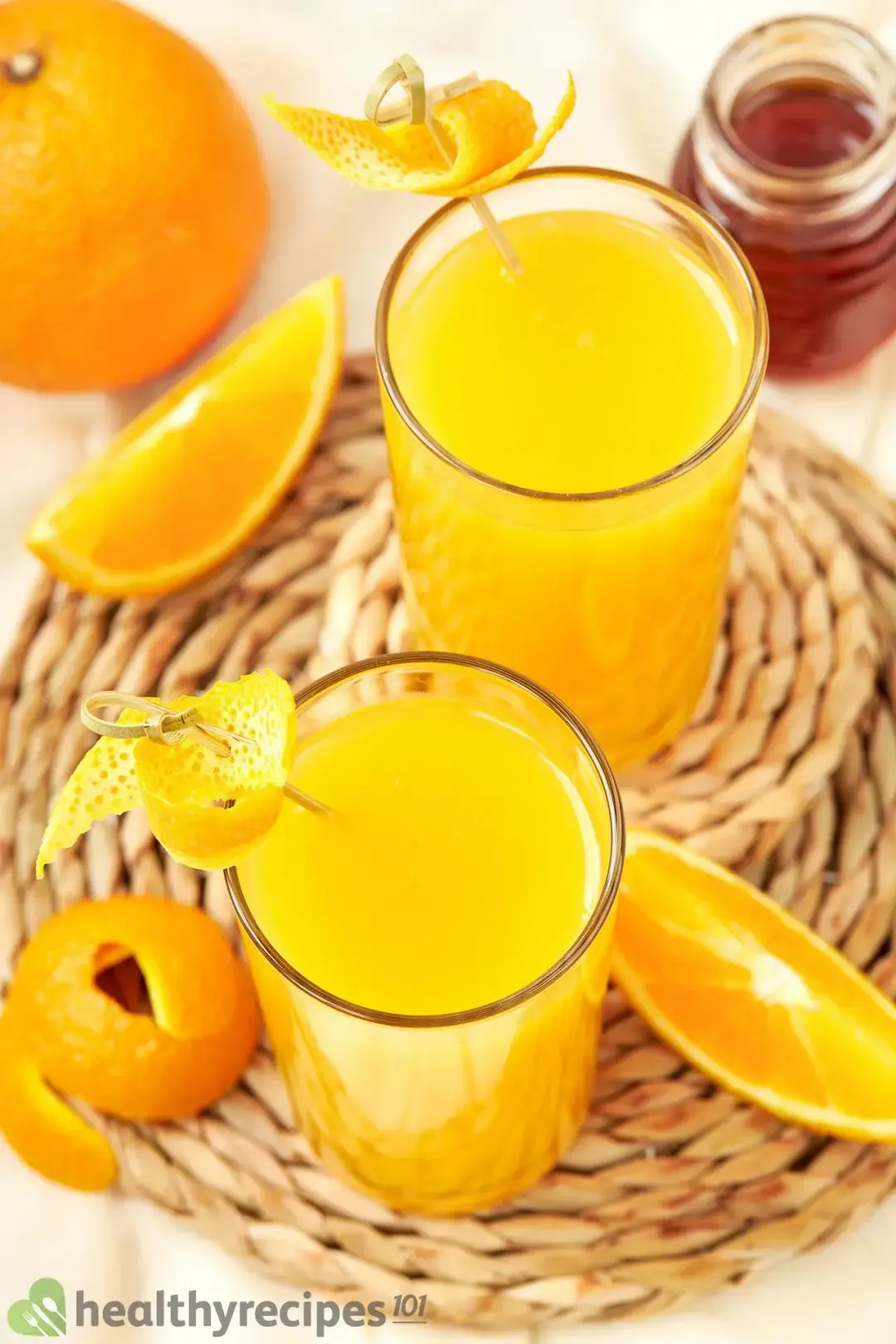 Two glasses of orange juice on a wood-colored coaster along with orange wedges