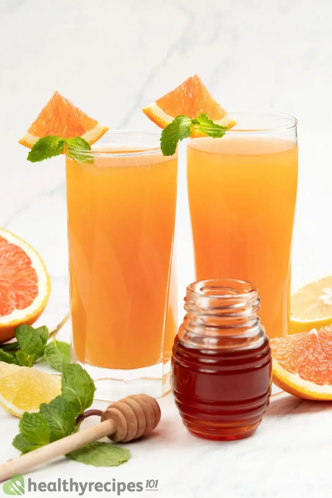Two glasses of grapefruit juice garnished with mints, grapefruit wedges all around, and a jar of honey