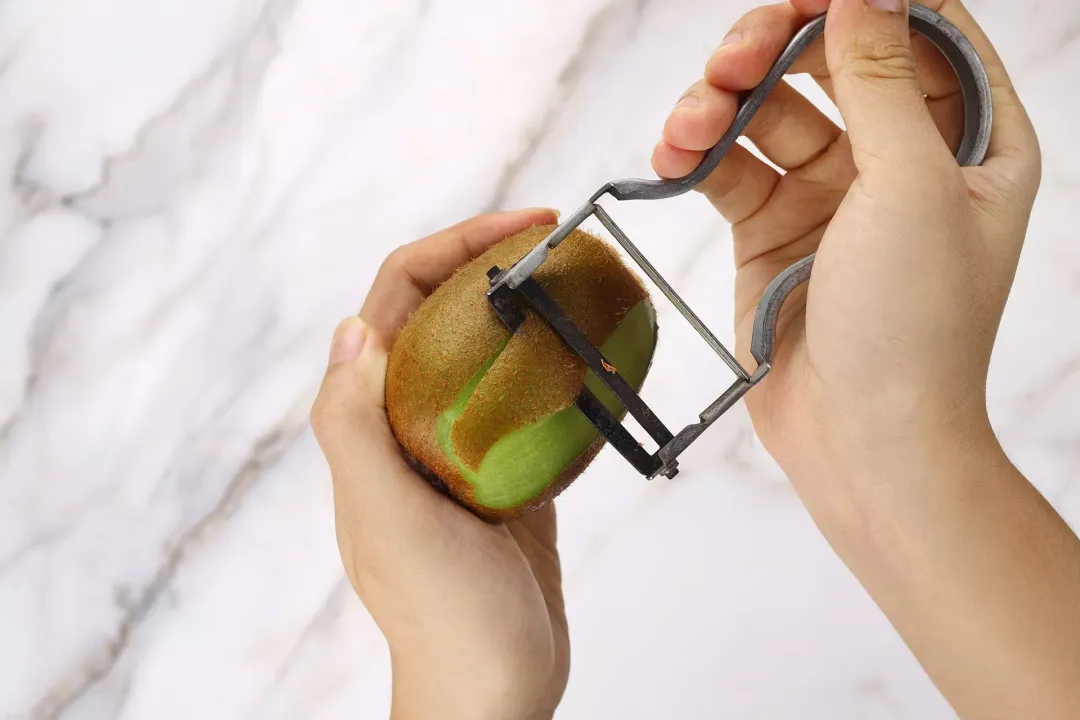 Two hands peeling a kiwi with a fruit peeler