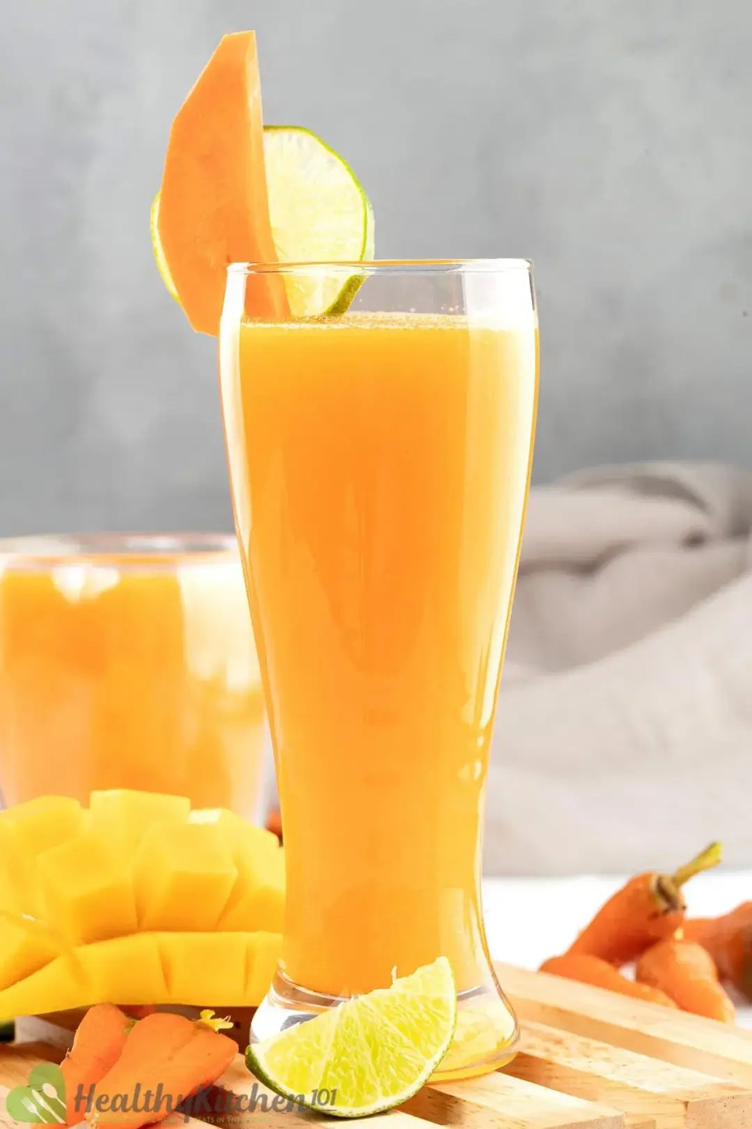 A tall glass of a mango smoothie garnished with a mango wedge, a lime wheel, and put next to lots of fruits