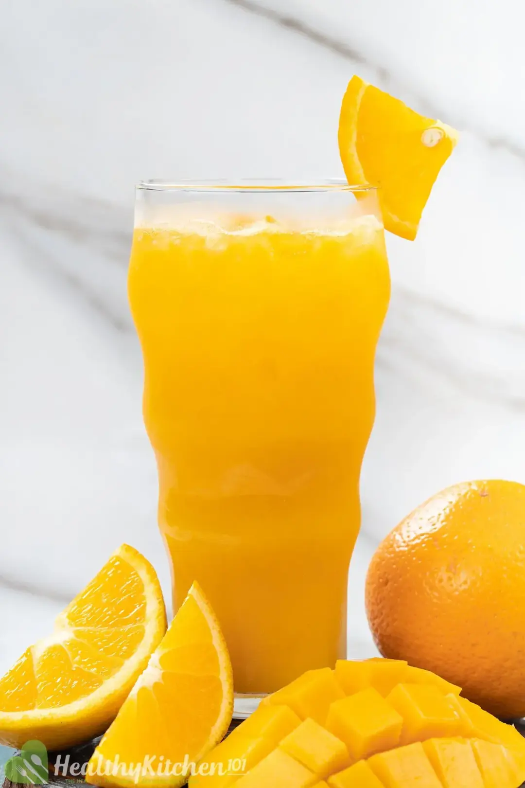 A tall glass of iced orange juice, next to some orange wedges, a whole orange, and a mango piece
