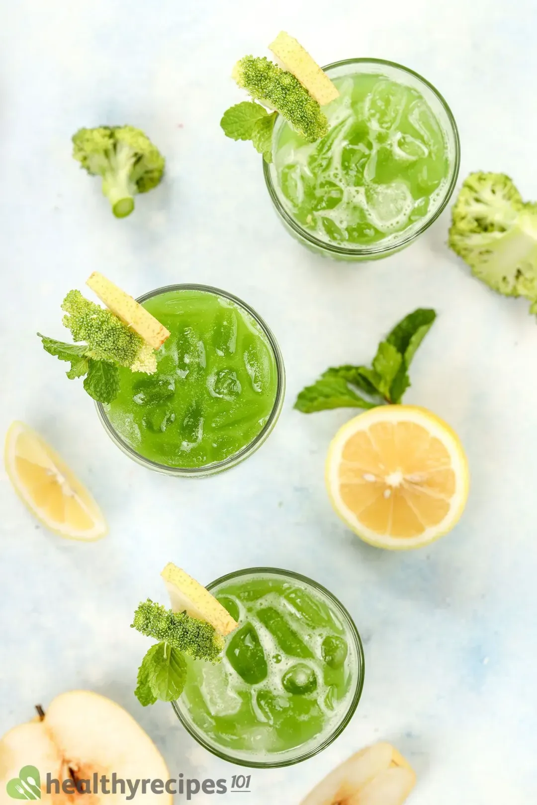 Three glasses of broccoli juice drinks taken from above, next to lime wedges, mints, and some apples