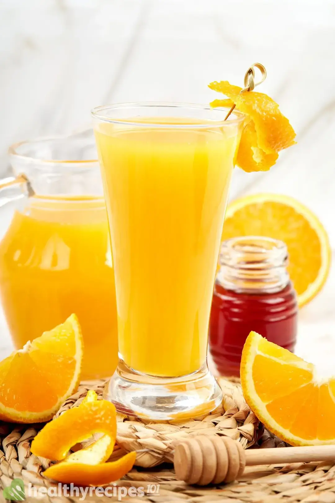 A glass of orange juice next to an orange juice pitcher, a honey jar, some orange wedges, and orange 