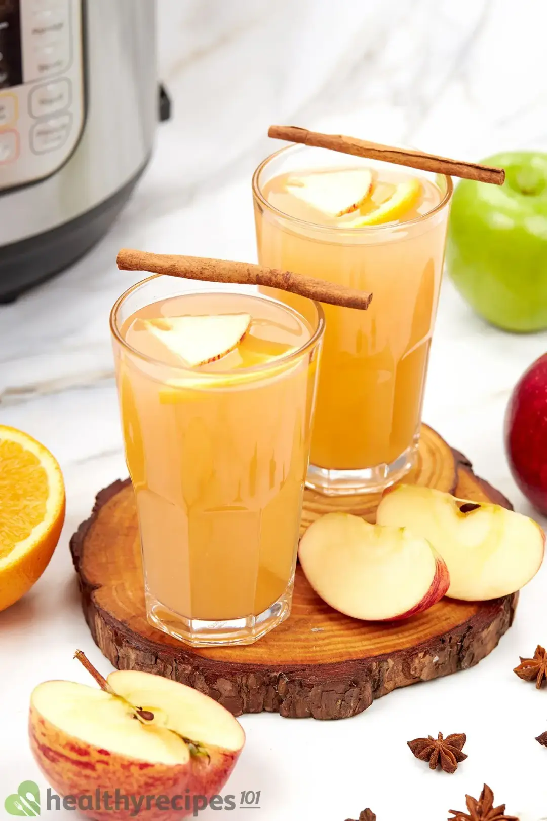 Two glasses of homemade apple cider, topped with cinnamon sticks and put next to lots of apple wedges and star anise