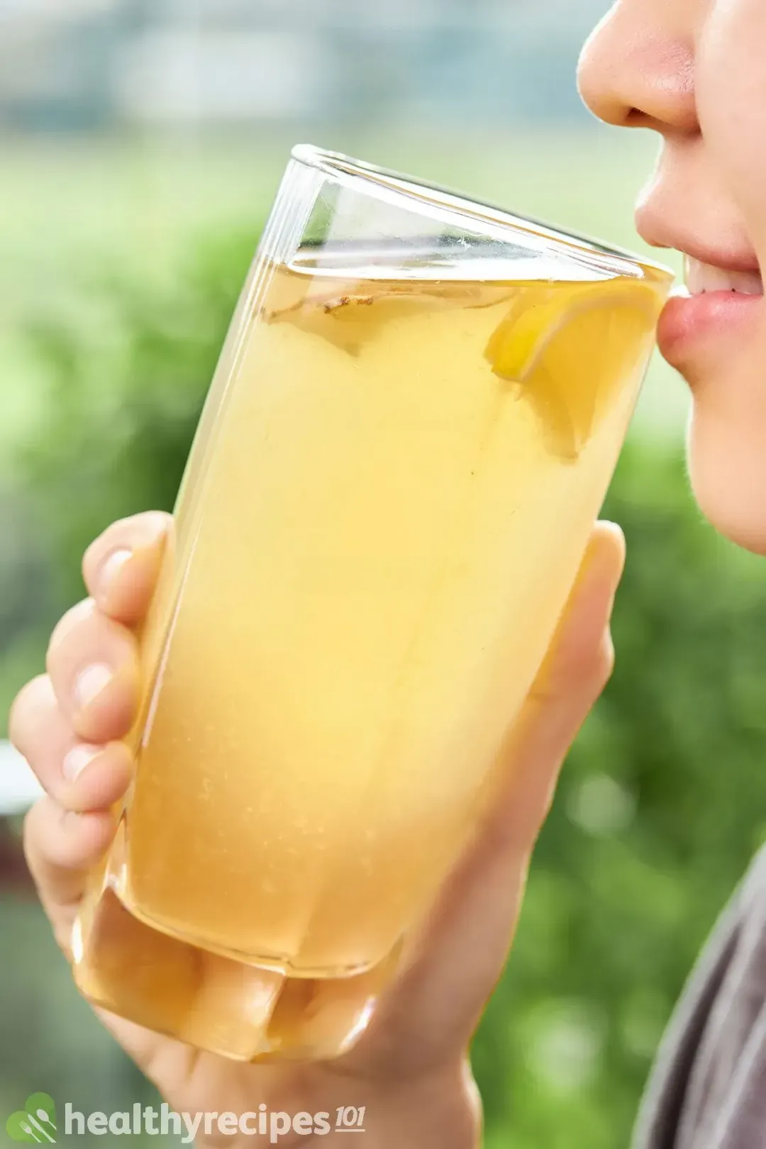 A tall glass filled with ginger and lemon honey vinegar drink held close to a mouth, about to be drunk