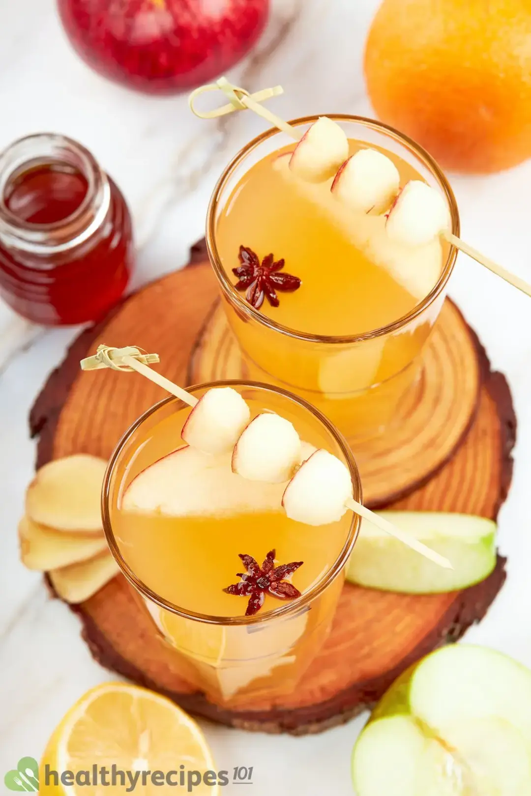 Two glasses of hot toddy cocktail garnished with apple wedges, apple skewers, star anise, and a jar of honey next to them