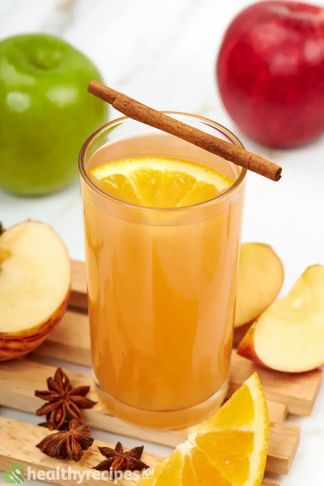 A tall glass of apple cider, with a cinnamon stick on top and lots of fruit wedges around