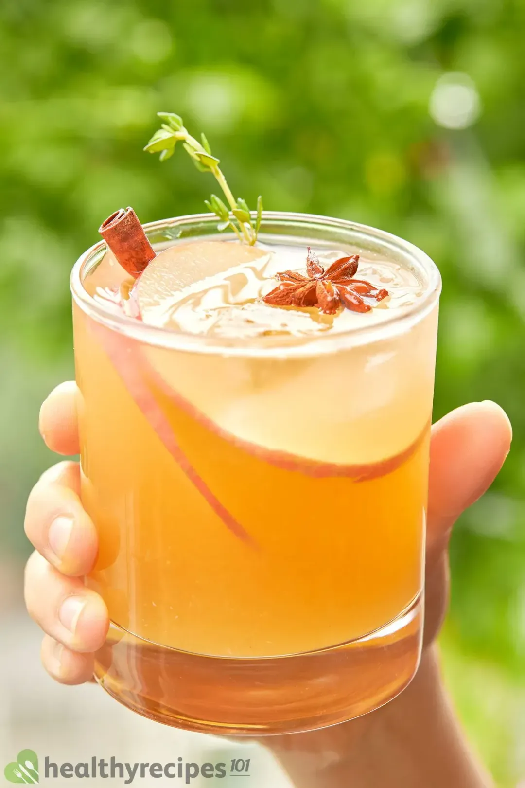 A hand holding a glass of apple cider cocktail, filled with a rosemary sprig, a star anise, and apple slices