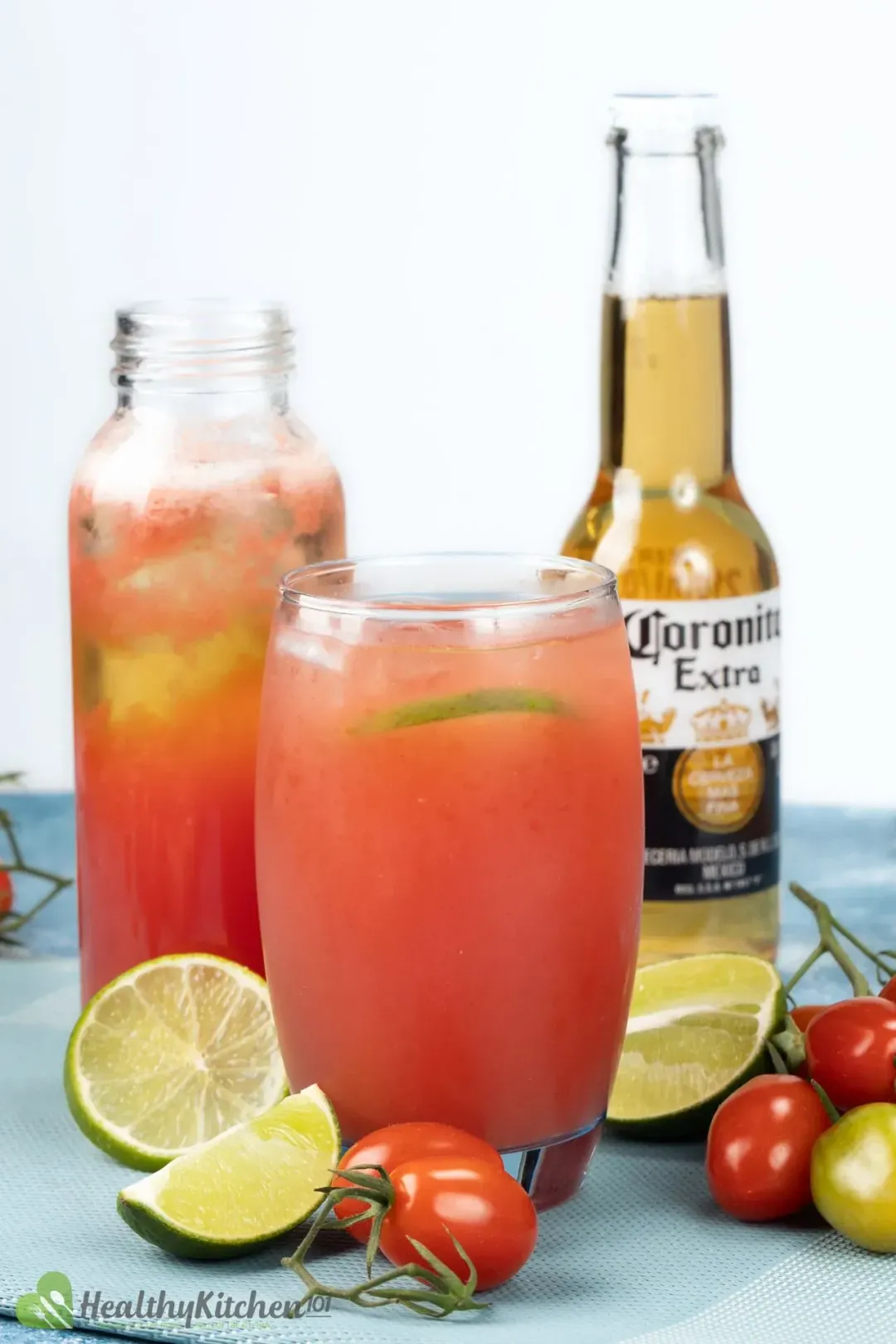 A chilled tomato and beer cocktail next to a bottle of red juice, beer bottle, lime wheels and cherry tomatoes