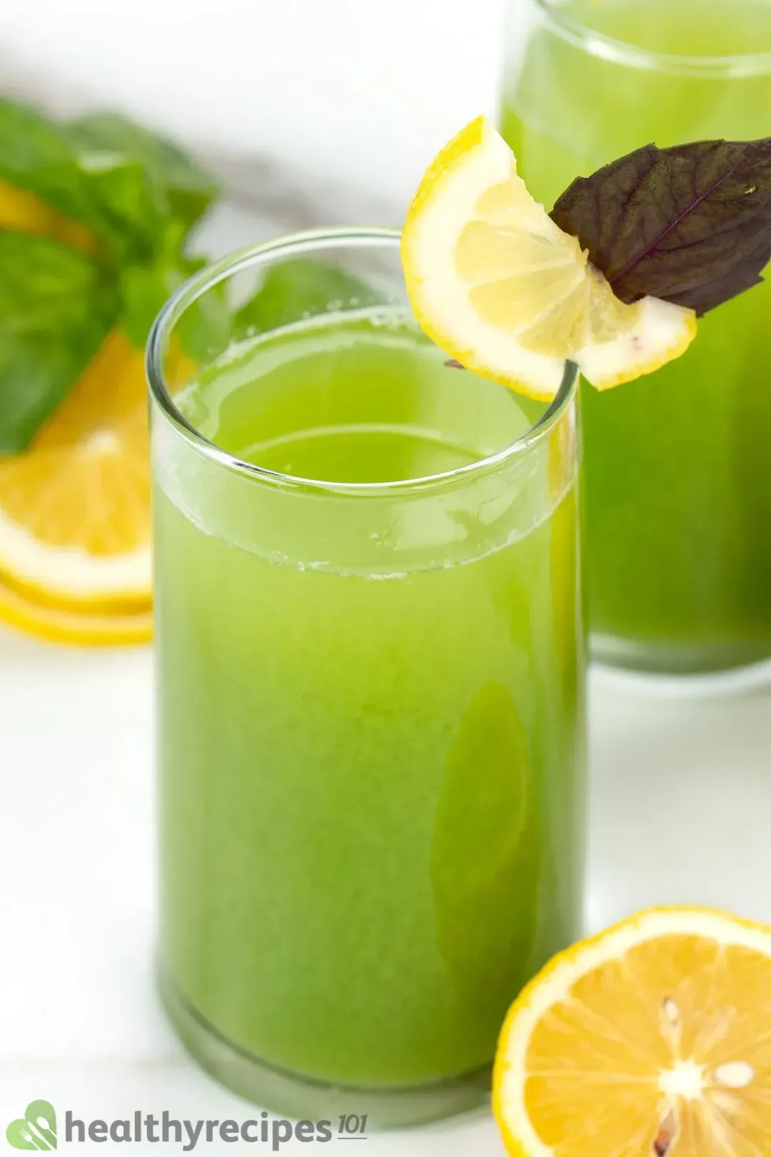 A glass of cucumber celery drink, with a lemon wedge and purple leaf on the rim, next to some lemon wheels and mints