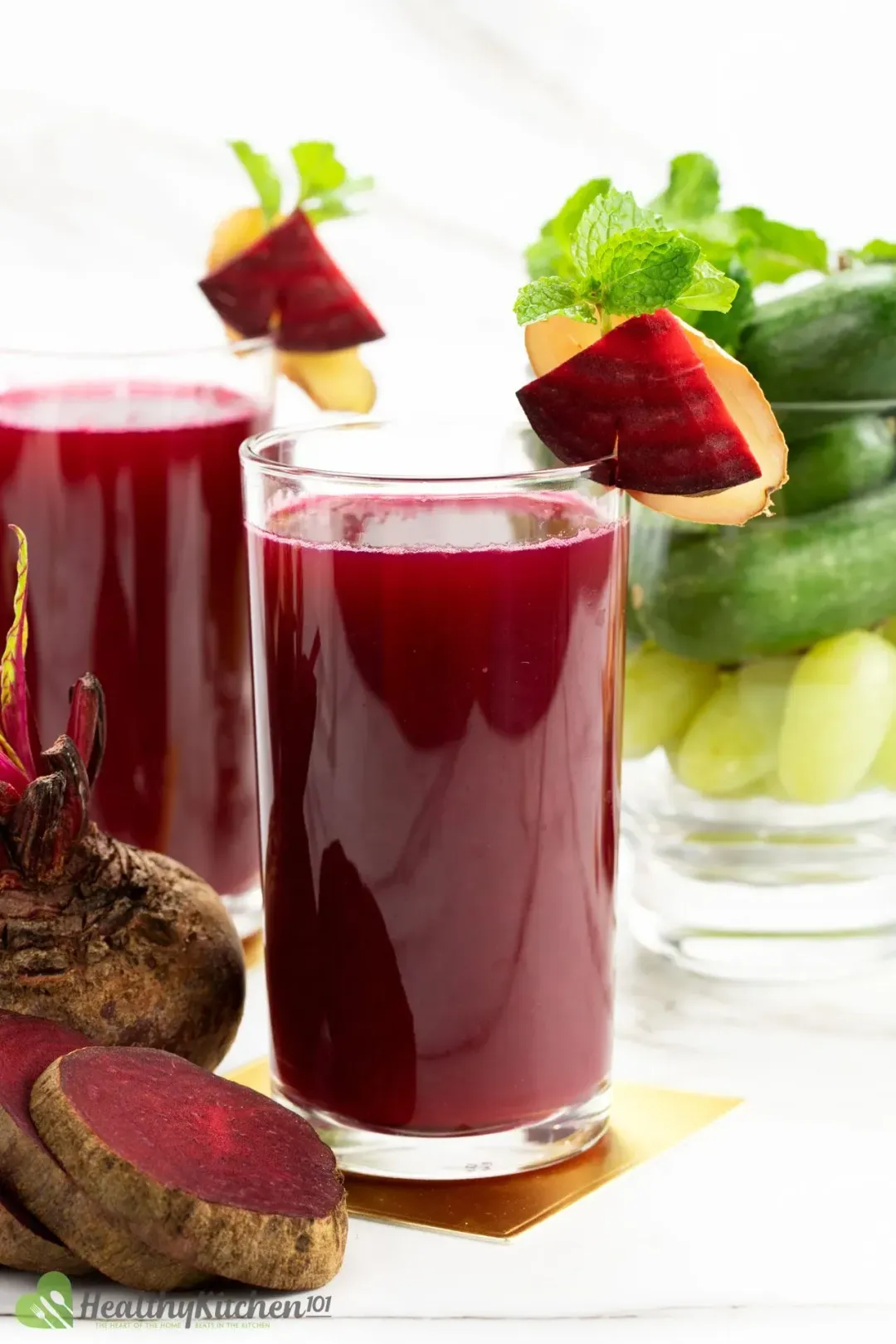 Two glasses of beetroot juice with beetroot and ginger pieces on the rim, next to sliced beetroots and cucumbers