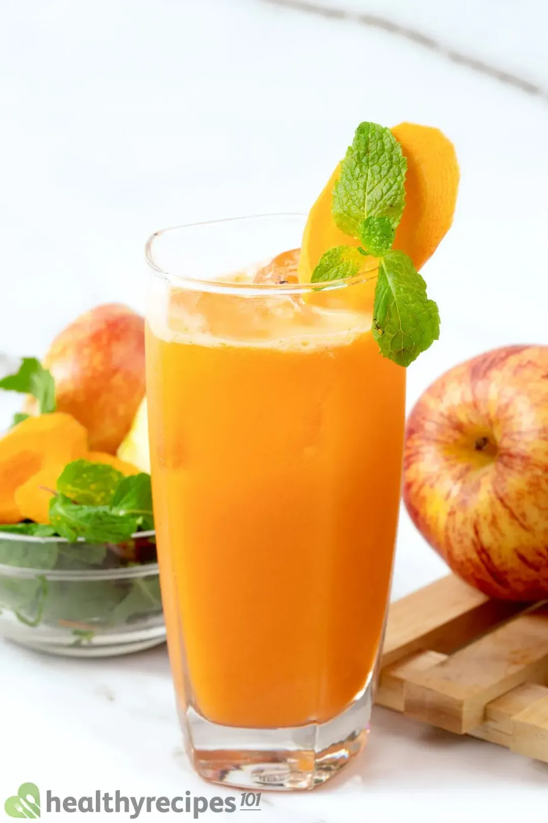 A tall glass of Orange drink with ice garnished with mints, carrot slices and whole apples