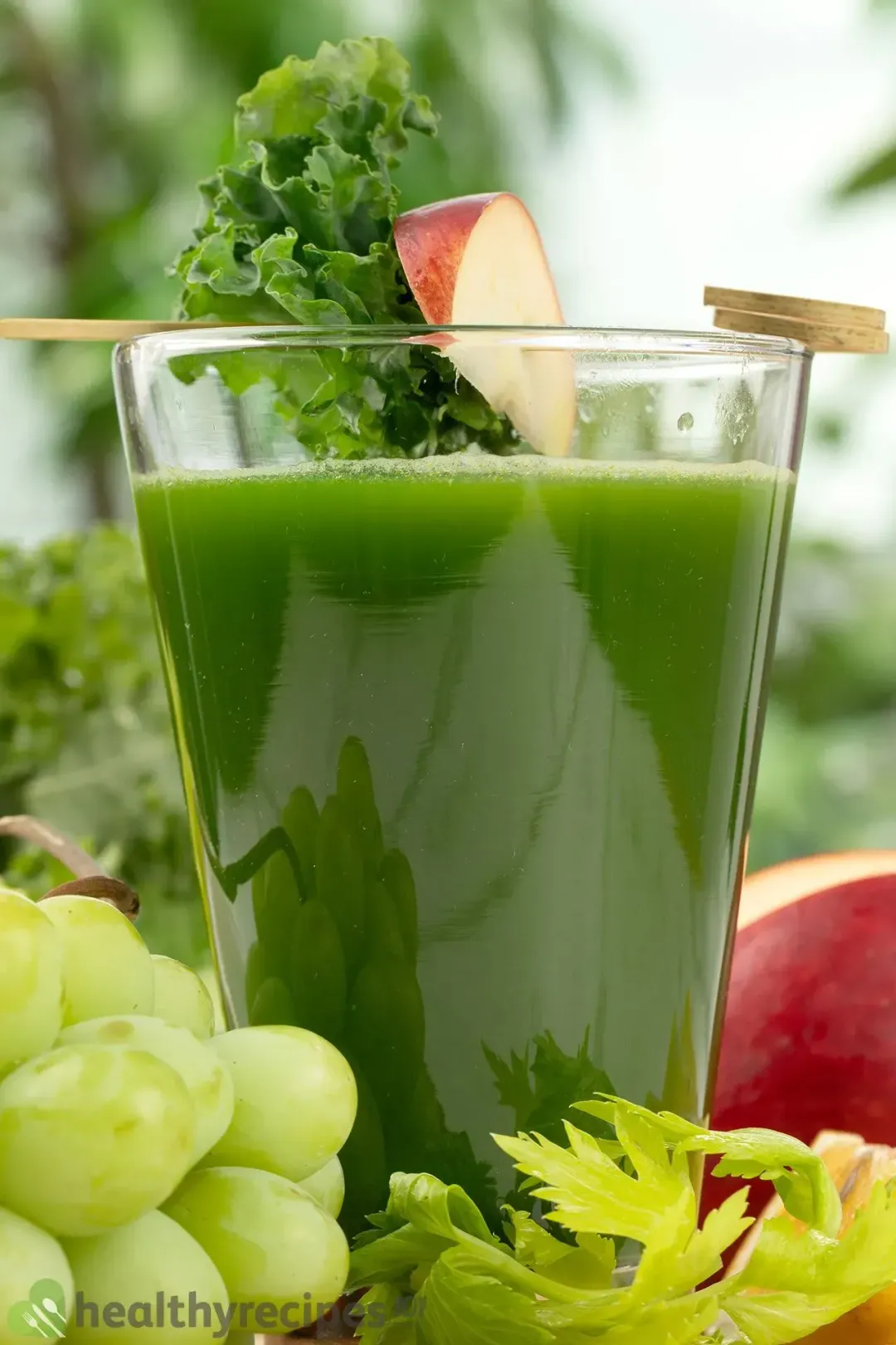  A close-up shot of a glass of green machine juice with some green grapes, kale leaves, celery, and an apple on the side for decorations