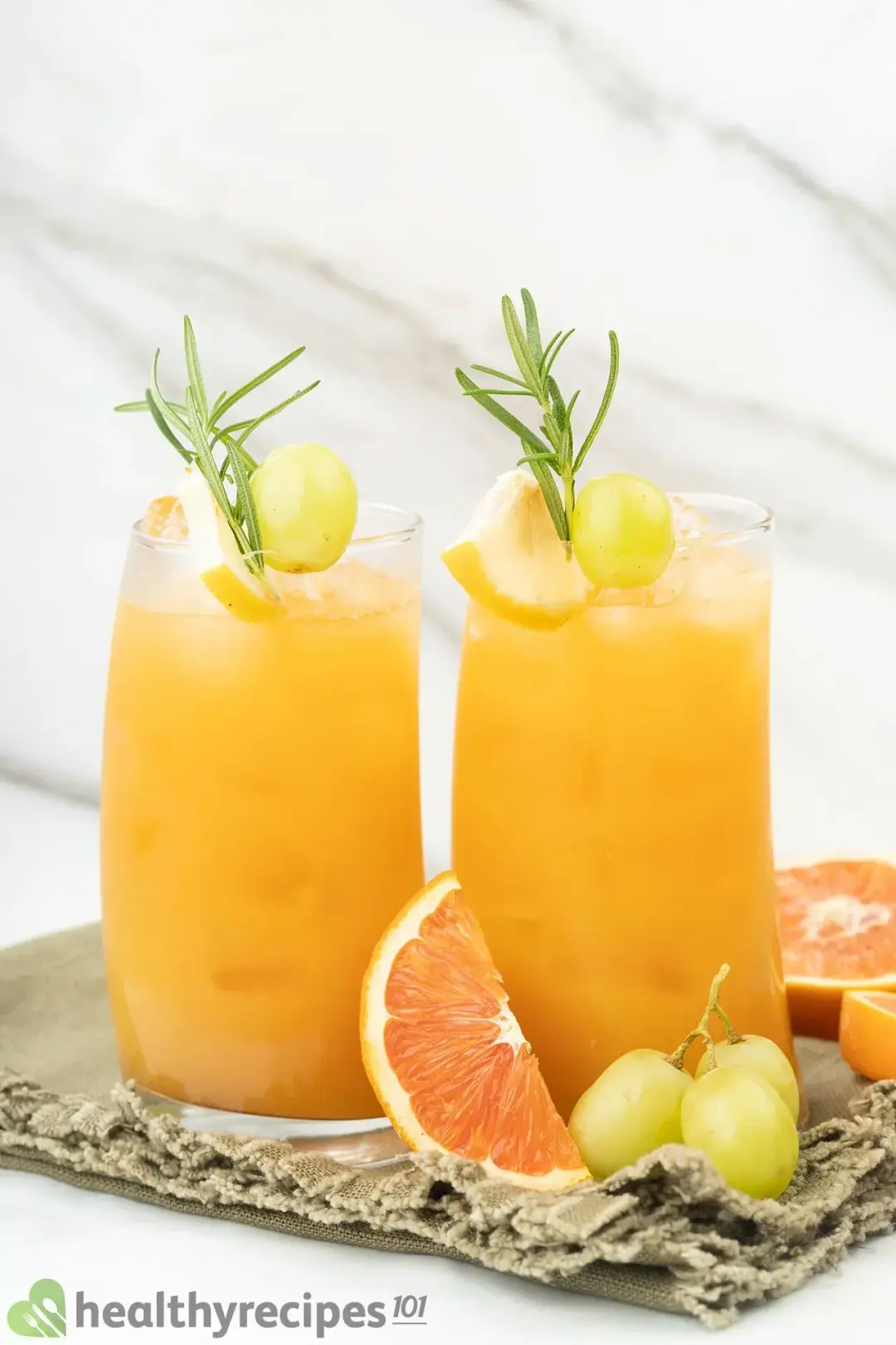 Two tall glasses of iced grapefruit juice each garnished with green grapes, rosemary sprigs, and put next to grapefruits and grapes