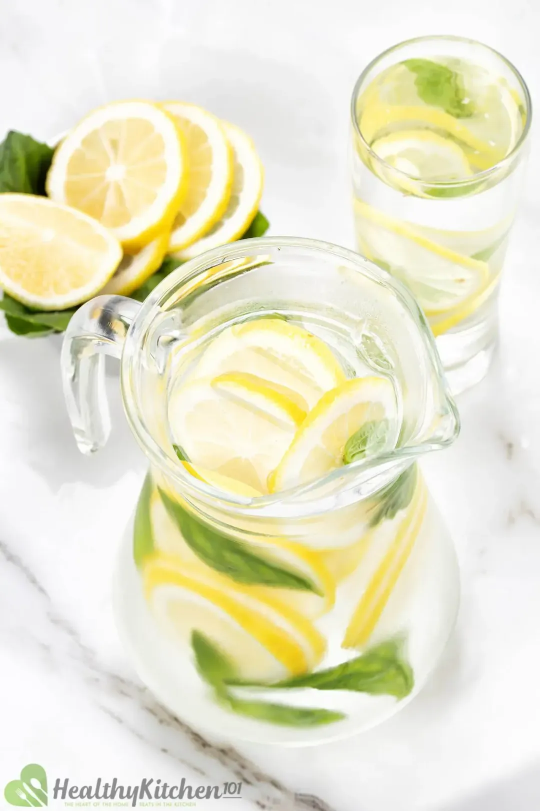 A pitcher and a glass of lemon and mint water next to a bowl of lemon wheels on some basils