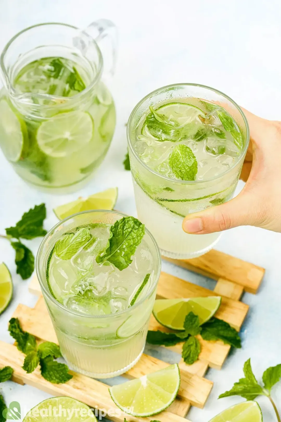 Two glasses of mojitos and a cute pitcher of mojitos, with mints and lime wedges lying around