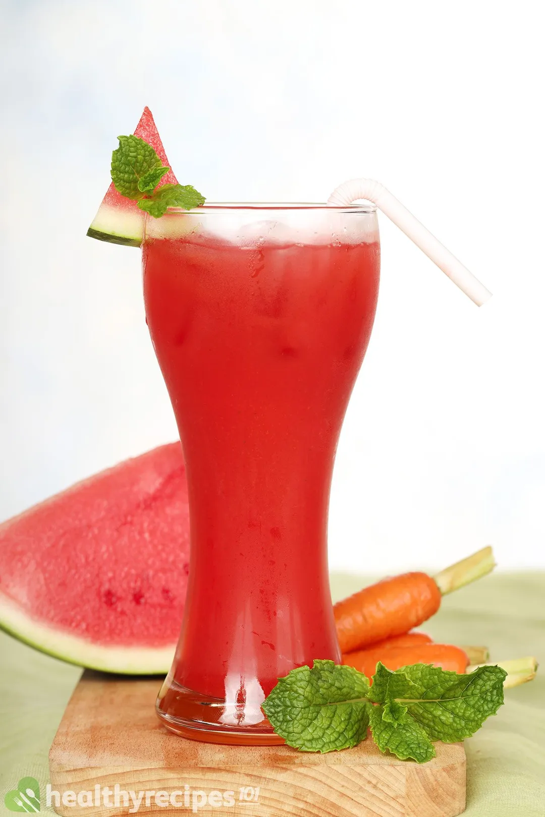 A tall glass of watermelon carrot juice placed on a wooden board with mint leaves, carrots, and a watermelon slice.
