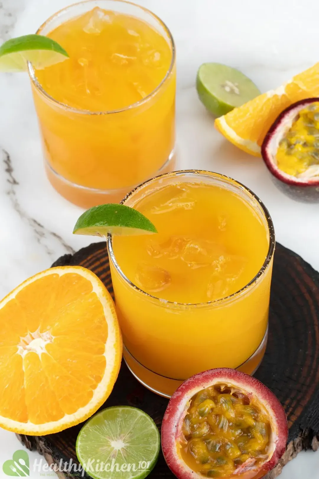 Two glasses of passion fruit margarita cocktails with lime wedges, next to halved passion fruit, lime, and orange